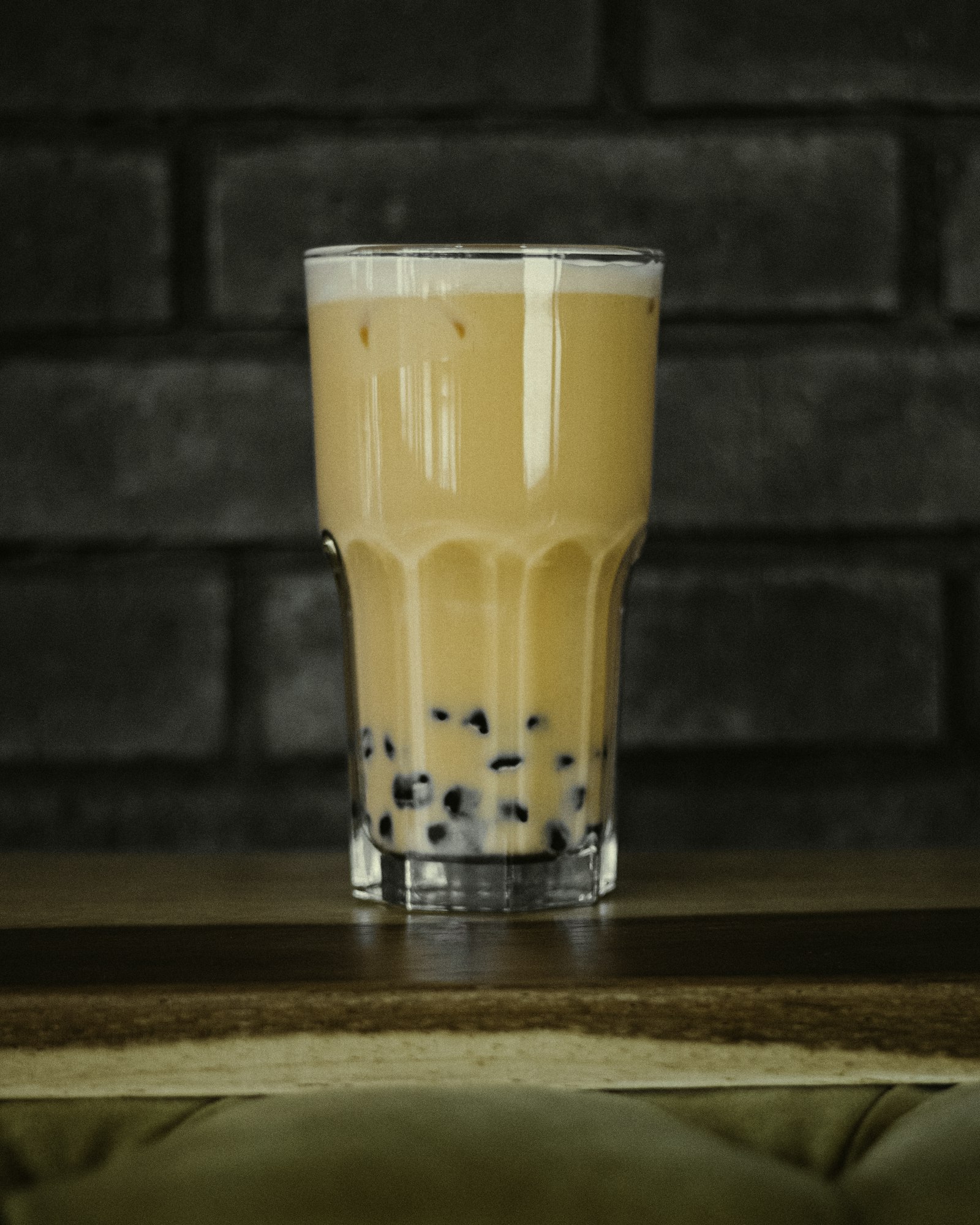 A tall glass of milk tea with tapioca pearls at the bottom in front of a brick wall
